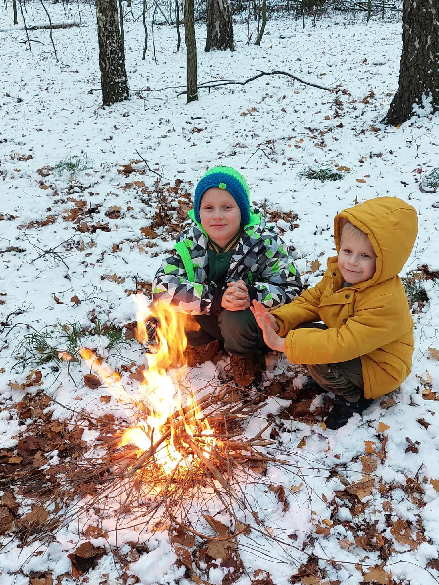 Mężczyzna w zimowej odzieży operuje przy ognisku rozpalonym na śniegu w nocy. W tle widać inne osoby, w tym jedną w białej kurtce odblaskowej, oraz znaki turystyczne na słupku.
